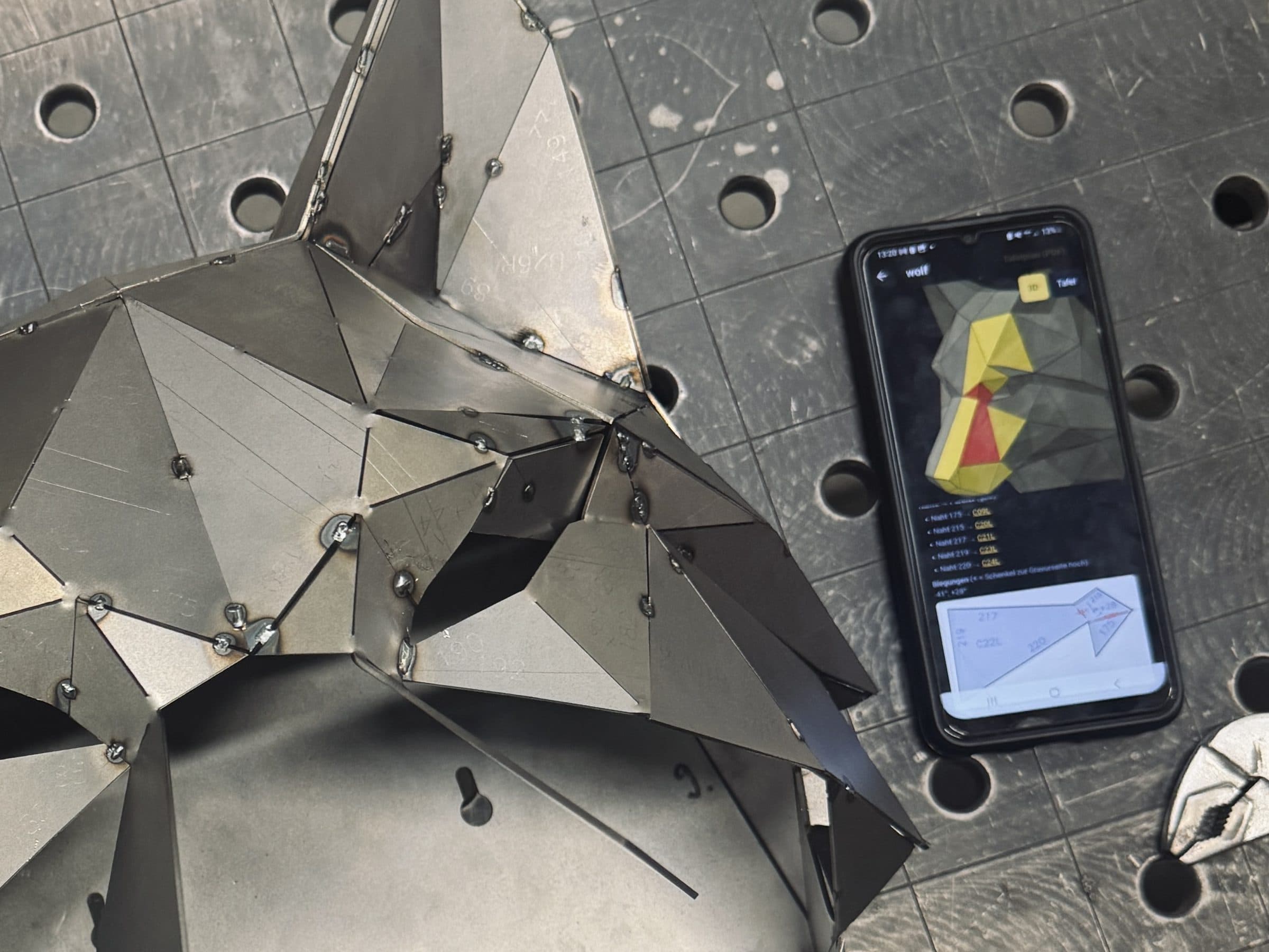 Handy mit der Montage-App neben einem angefangenen Wolfskopf auf dem Schweißtisch